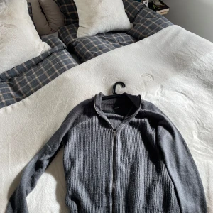 Mörkgrå stickad kofta med dragkedja - Säljer en mörkgrå stickad kofta från OCHNIK med hel dragkedja framtill. Koftan har ribbad struktur, hög krage och långa ärmar. Perfekt för lager-på-lager och enkel att matcha med olika outfits.