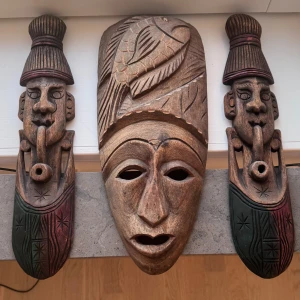 Afrikansk mask - Bilderna visar tre handgjorda träskulpturer/masker med etniska motiv och färgdetaljer. De är konstföremål, inte elektronik. Inga synliga elektroniska komponenter, märken eller modeller. Skicket ser bra ut med naturligt slitage från träarbetet.