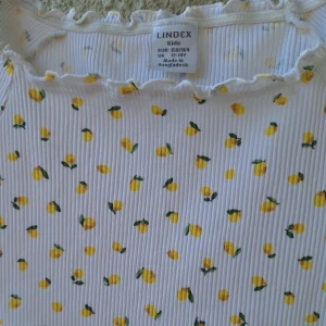 Vit ribbad topp med citronmönster Lindex - Superfin vit ribbad topp från Lindex med sött citronmönster i gult och grönt. Toppen har korta ärmar, volangkant vid halsringning och ärmslut samt en figurnära passform. Perfekt för en fräsch och somrig look! Skicka gärna ett bud på pris så kan vi komma överens,men just nu ligger den på 80kr. 