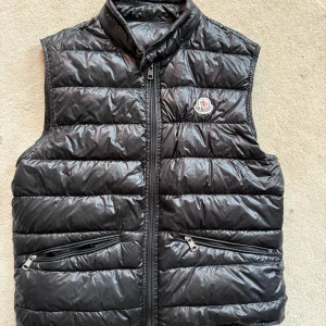 Svart ÄKTA Gui dunväst från Moncler - Snygg svart dunväst från Moncler med quiltad design och hög krage. Västen har två praktiska fickor med dragkedja framtill och Moncler-logga på bröstet. 100 procent äkta och i toppskick. Pris på Monclers hemsida 9260 strl 2 motsvarar s/m