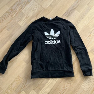 Svart Adidas sweatshirt med logga - Svart sweatshirt från Adidas med den klassiska vita trefoil-loggan och text framtill. Tröjan har rund halsringning och långa ärmar. Tillverkad i mjuk bomullsmix som är skön att ha på sig. Perfekt för en sportig och avslappnad stil.