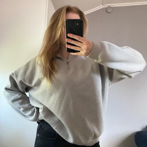 Grå half zip sweatshirt - Grå oversized sweatshirt med broderad logga på bröstet. Tröjan har half zip-dragkedja, ribbade muddar och lång ärm. Perfekt för chill dagar och enkel att styla med jeans eller leggings.