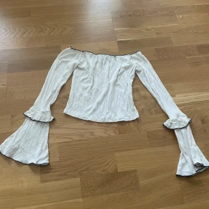 Vit offshoulder topp med volangärmar - Snygg vit offshoulder topp med diskreta ränder och långa trumpetärmar med volangdetaljer och svarta kanter. Toppen har en figurnära passform och är perfekt för dig som vill sticka ut med en trendig look.