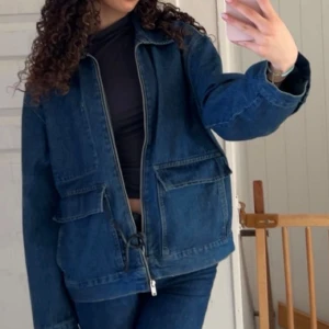 Oversized jeansjacka med dragkedja - Säljer en oversized jeansjacka i mörkblå denim med dragkedja framtill och stora fickor på båda sidor. Jackan har klassisk krage och långa ärmar med knapp vid ärmslut. Perfekt för dig som gillar en chill och avslappnad stil.