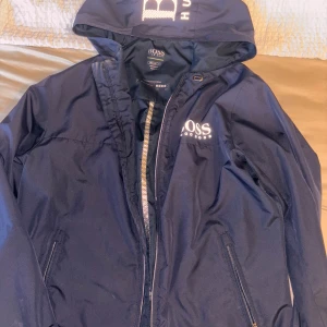 Marinblå vindjacka Hugo Boss - Snygg marinblå vindjacka från Hugo Boss med huva och dragkedja. Jackan har vita logotryck på bröstet och huvan, samt praktiska fickor med dragkedja. Perfekt för blåsiga dagar.