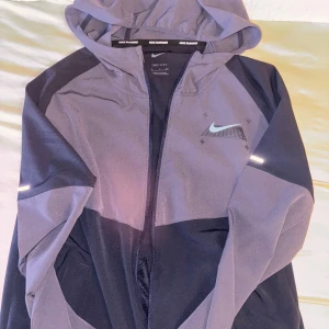 Nike Running vindjacka med huva - Nike Running vindjacka i mörkblå och ljusare blå/lilla med reflekterande detaljer och hel dragkedja. Jackan har huva, lång ärm och sportig design med Drifit-material som andas. Perfekt för löpning och utomhusträning.