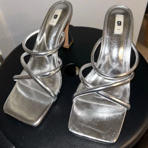 Silvriga sandaler med klack från Gina Tricot - Snygga sandaler i metallic silver med fyrkantig tå och höga klackar. Skorna har tunna remmar i syntetmaterial som korsar över foten och ger en trendig look. Perfekta för att lyfta vilken outfit som helst. Använda en gång därav topp skick. True to size. Ny pris: 499kr.