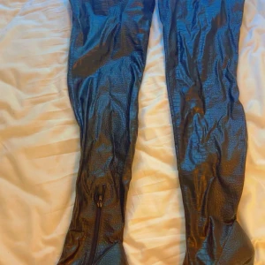 Svarta höga stövlar med krokoprägling - Säljer ett par svarta höga stövlar med krokodilpräglat mönster och dragkedja på insidan. Snyggt glansigt material som ger en edgy look. Perfekta att styla till kjol eller över jeans för en statement-outfit.