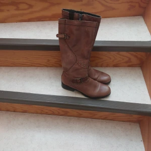 Bruna höga boots med spännen - Snygga bruna boots i skinn med dekorativa spännen och nitar. De har rund tå, platt sula och dragkedja på insidan. Insidan är fodrad med ett mönstrat tyg i fiskbensmönster. Perfekta för dig som vill ha en cool och tidlös stil.