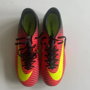 Nike Mercurial fotbollsskor röd/svart - Säljer ett par Nike Mercurial fotbollsskor i storlek 40. Skorna är röda med svarta ränder och har en stor gul Nike-logga på sidan. De har snörning och dobbar i gult för bra grepp på planen. Perfekta för dig som vill sticka ut på fotbollsplanen.