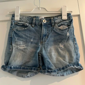 Blå jeansshorts med slitningar 157 - Snygga blå jeansshorts från 157 i storlek 160. De har coola slitningar framtill, klassiska fem fickor och uppvikta benslut. 