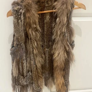 Brun pälsväst med fluffig look - Säljer en brun pälsväst med naturliga färgskiftningar och lång, fluffig päls. Västen har en cool, bohemisk vibe och är perfekt för att lyfta vilken outfit som helst. Den är ärmlös och har en öppen front utan knäppning.