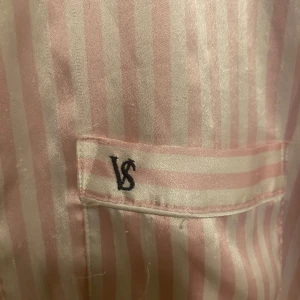 Randig rosa blus från Victoria's Secret - Säljer en lyxig blus från Victoria's Secret i glansigt material med rosa och vita ränder. Klassisk krage, knäppning framtill och bröstficka med VS-broderi. Säljs då jag inte använder den längre och måste rensa min garderob💞. 