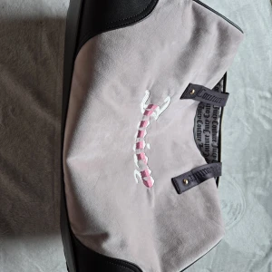 Juicy Couture weekend bag  - Stor och mjuk axelväska från Juicy Couture i ljuslila mocka med svarta detaljer och axelremmar med logga. Broderad Juicy-text i vitt och rosa på framsidan. Fodret är mönstrat med Juicy Couture-tryck. Perfekt för dig som gillar statement-väskor.