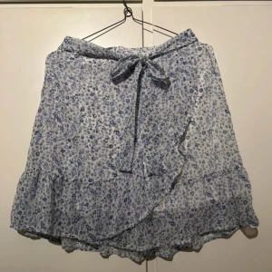 Blommig omlottkjol från Vero Moda - Supersöt kort sommarkjol från Vero Moda i vitt med blått blommönster. Kjolen har volangkant och knytband i midjan för extra detalj. Perfekt för en fräsch och somrig look.