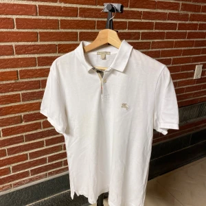 Vit Burberry pikétröja i bomull - Vit pikétröja från Burberry med klassisk krage, korta ärmar och broderad logga på bröstet. Tillverkad i mjuk bomull och har knäppning framtill med två knappar. Perfekt för en clean och stilren look.