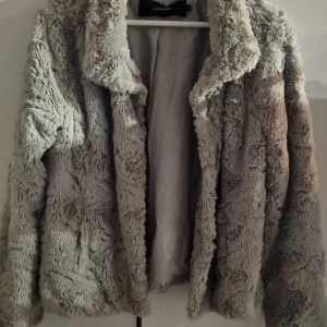 Grå teddyjacka från Vero Moda - Supermjuk och fluffig grå teddyjacka från Vero Moda i storlek M. Jackan har hög krage, långa ärmar och stängs med knappar framtill. Perfekt för dig som vill ha en mysig och trendig look under kyliga dagar.