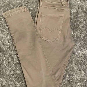 Beige Replay Anbass Hyperflex jeans herr - Snygga beige Replay Anbass Hyperflex jeans och diskreta sömdetaljer på bakfickorna. Tillverkade i mjuk bomullsblandning med stretch för extra komfort. Perfekta för en stilren och avslappnad look.