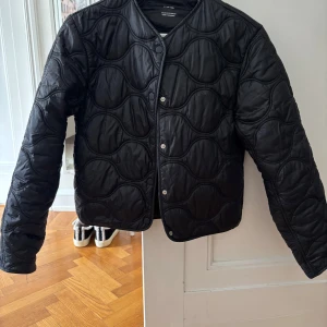 Mango jacka - Säljer en svart quiltad jacka från Mango med vågformat mönster. Jackan har tryckknappar framtill, rundad hals och lång ärm. Materialet är glansigt och känns lätt, perfekt för lager-på-lager under höst och vår.