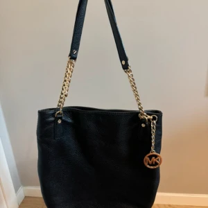 Svart axelväska Michael Kors i skinn - Snygg svart axelväska från Michael Kors i mjukt skinn med guldfärgade kedjedetaljer och MK-hänge. Väskan har både axelrem och handtag, samt flera innerfack med logotypmönstrat foder. Perfekt för dig som vill ha en stilren och rymlig väska med lyxig känsla.