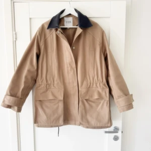 Beige parkajacka från COS med svart krage - Stilren beige parkajacka från COS med  skinnkrage. Jackan har stora fickor framtill, dold dragkedja och tryckknappar, samt justerbar midja. Tillverkad i bomullsmix med en loose passform och är over size.