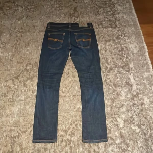 Mörkblå jeans från Nudie Jeans - Snygga mörkblå jeans från Nudie Jeans med klassisk orange söm och vågig detalj på bakfickorna. Jeansen har rak passform och är tillverkade i slitstarkt denim. Perfekta för dig som gillar stilrena och tidlösa jeans med en cool touch.