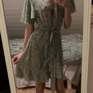 Grön blommig omlottklänning med knyt - Supersöt grön kort omlottklänning med vitt blommönster och knyt i midjan. Klänningen har v-ringning, volangiga korta ärmar och luftig passform. Perfekt för varma dagar och ger en fräsch och somrig vibe.