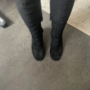 Svarta höga stövlar i mocka - Säljer ett par svarta höga stövlar med klack och rund tå. Stövlarna är i mjuk mocka och har dragkedja på sidan för enkel påtagning. Perfekta för att lyfta din outfit med en edgy vibe.