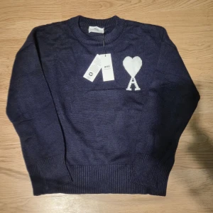 Mörkblå stickad tröja från Ami - Snygg mörkblå stickad tröja från Ami med vit hjärtlogga och bokstaven A på bröstet. Klassisk rund halsringning och ribbade muddar vid ärmslut och nederkant. Perfekt för dig som gillar stilrena och trendiga plagg.