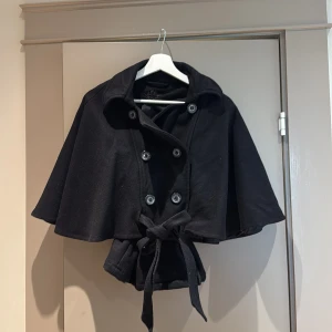  poncho kappa svart - Stilren svart kappa med kort, vid passform och knytband i midjan. Dubbelknäppt framtill med stora knappar och bred krage. Perfekt för dig som vill ha en snygg och unik jacka med cape-vibbar.