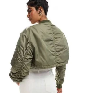 Croppad grön bomberjacka - Snygg croppad bomberjacka i olivgrönt med klassisk ribbad krage och muddar. Jackan har lång ärm, dragkedja framtill och en liten ficka på bröstet. Materialet är glansigt och känns lätt, perfekt för att lyfta din streetstyle.