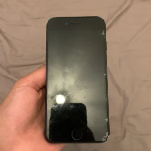 iPhone 7 - Begagnad iPhone 7 i svart. Skärmen har flera sprickor och repor, särskilt i nedre hörnet. Baksidan och kanterna visar tydligt slitage och repor. Touch ID-knappen syns tydligt. Perfekt som reservtelefon eller för den händige som kan byta skärm.