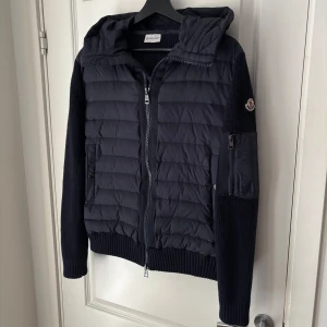Mörkblå Moncler Cardigan - Snygg mörkblå cardigan från Moncler med stickade ärmar och huva. Jackan har quiltad front, dragkedja hela vägen och praktiska fickor med knappar. Ärmarna är ribbstickade och det finns en extra ficka med dragkedja på ena ärmen. Perfekt nu till hösten. 