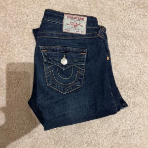 True Religion mörkblå bootcut jeans - Mörkblå jeans från True Religion med klassiska kontrastsömmar och snygga fickdetaljer med silverknappar. Modellen har bootcut-ben och låg midja. **((Dem är uppsydda för personer under 160))**🌸