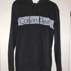 Svart Calvin Klein hoodie med logga - Svart hoodie från Calvin Klein Jeans med stor grå logga över bröstet. Klassisk känguruficka framtill, huva med snörning och långa ärmar. Perfekt för en chill och stilren look.