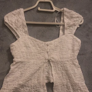 Vit linnetopp med knappar och struktur - Supersöt vit linnetopp med breda axelband och snygg struktur i tyget. Toppen har dekorativa knappar framtill och en öppen detalj nedtill som ger en cool look. Perfekt för varma dagar och enkel att styla med jeans eller kjol.