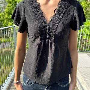 Svart blus med spets och v-ringning - Säljer en svart blus med v-ringad hals och fina spetsdetaljer framtill. Blusen har korta, lite fladdriga ärmar och ett diskret hålmönster i tyget. Passformen är normal och materialet känns lätt och luftigt, perfekt för varmare dagar.