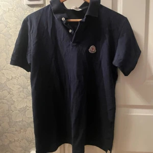 Mörkblå Moncler piké t-shirt - Snygg mörkblå piké t-shirt från Moncler med klassisk krage och två vita knappar framtill. T-shirten har korta ärmar och Moncler-logga på bröstet. Perfekt för dig som gillar stilrena och exklusiva plagg.