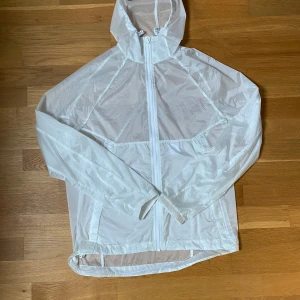 Vit vindjacka från J.Lindeberg - Säljer en tunn och lätt vit vindjacka från J.Lindeberg. Jackan har huva, hel dragkedja framtill och är tillverkad i ett glansigt syntetmaterial. Perfekt för blåsiga dagar och enkel att packa ner i väskan. Snygg och stilren design med diskret logga på bröstet.