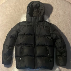 Svart dunjacka från Tommy Hilfiger - Säljer en svart pufferjacka från Tommy Hilfiger med huva och dragkedja. Perfekt för kalla höst och vinter dagar! 