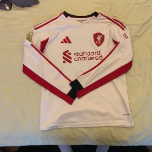 Liverpool långärmad bortatröja Adidas - Liverpool FC långärmad bortatröja från Adidas med tryck 'Wirtz 7' på ryggen. Tröjan är vit med röda detaljer, klubbmärke och sponsortryck framtill. Svart krage och röda ränder längs ärmarna. Tröjan är i nyskick 