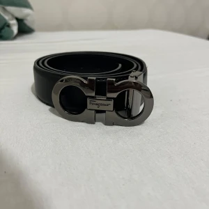 Svart bälte Ferragamo - Snyggt svart bälte från Salvatore Ferragamo med ikoniskt metallspänne i silverfärg. Bältet är tillverkat i slätt skinn och har en stilren, klassisk design som lyfter vilken outfit som helst. Perfekt för dig som gillar exklusiva accessoarer och vill sticka ut med detaljer.