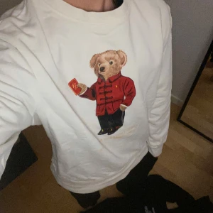 Vit tröja med Polo Bear från Ralph Lauren - Vit långärmad sweatshirt från Ralph Lauren med ikoniska Polo Bear-trycket framtill. Björnen har röd jacka och håller ett rött kort. Tröjan har rund halsringning och är i mjuk bomull. Perfekt för dig som gillar streetstyle och klassiska märken.