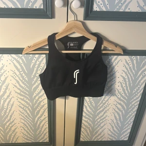 Svart sporttopp från RS Sports - Sportig svart topp från RS Sports med breda axelband och stilren logga framtill. Tillverkad i stretchigt material med 92% nylon och 8% spandex för en bekväm passform. Perfekt för träning och aktiva dagar.