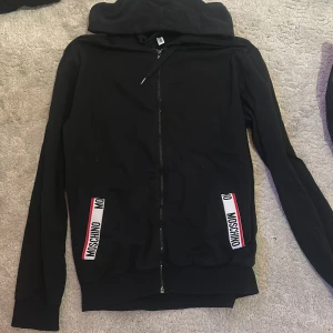 Svart hoodie från Moschino med dragkedja - Svart hoodie från Moschino med huva och hel dragkedja framtill. Snygga vita och röda Moschino-detaljer vid fickorna. Långärmad modell i mjuk bomull, perfekt för en chill och stilren look.