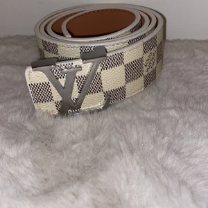 Vit och beige Louis Vuitton bälte - Säljer ett snyggt Louis Vuitton bälte i klassiskt damier-mönster med vita och beige rutor. Bältet har en silverfärgad LV-spänne och är gjort i skinn. Perfekt accessoar för att lyfta din outfit med en lyxig touch.