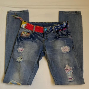 Desigual jeans med färgglada detaljer - Blå jeans från Desigual med straight fit, färgglada tygdetaljer i midjan och fickor, samt slitna och lagade partier med broderade sömmar. Strl 34.