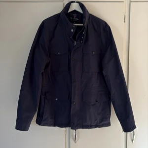 Mörkblå fieldjacket från Bondelid - Säljer en mörkblå fieldjacket från Bondelid med hög krage och fyra stora fickor framtill. Jackan har knappar och dragkedja framtill samt en stilren, minimalistisk look. Perfekt att matcha med olika outfits. Nypris: 1799 kr