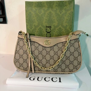 Ophidia  i klassiskt GG  - Snygg Ophidia  i klassiskt GG Supreme-mönster. Väskan är beige och med detaljer i beige skinn, guldfärgad kedja och dragkedja. Ikonisk GG-logga framtill och innerfack i ljust tyg. Perfekt för dig som vill ha en lyxig och trendig accessoar. Skickas snabbt med kartong och dustbag. 