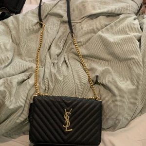 Svart YSL axelväska i skinn med kedja - Säljer en svart axelväska från YSL i quiltat skinn med guldiga detaljer och kedjerem.  Väskan har klassisk YSL-logga framtill, magnetlås och flera fack inuti. Perfekt storlek för det viktigaste och ger en lyxig vibe till din outfit.
